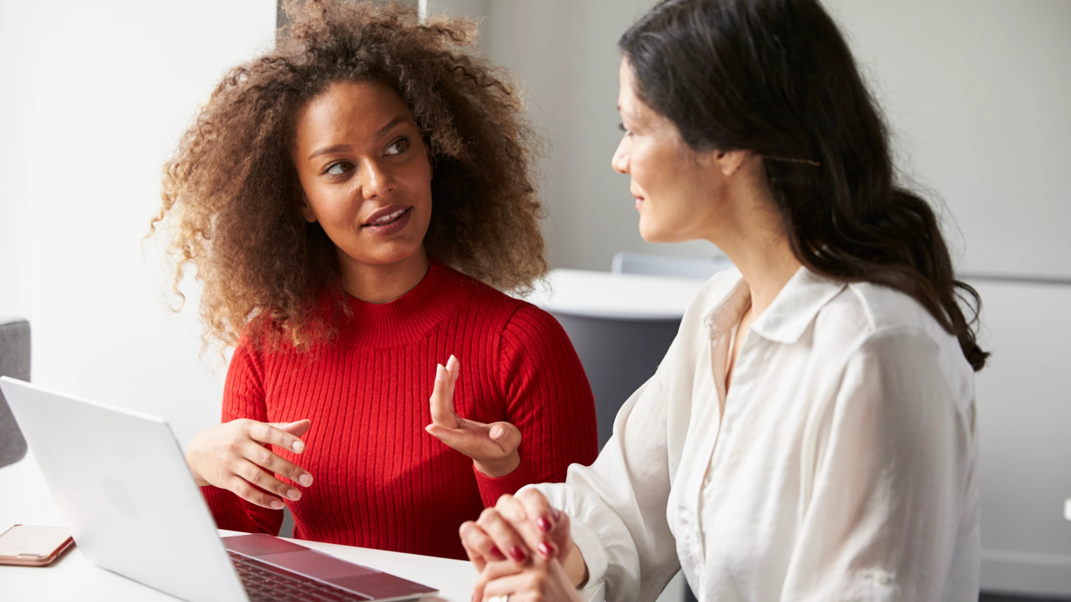 Una mujer negra con suéter rojo le explica algo a una nueva empleada: mujer blanca con blusa blanca. Palabras clave: cómo causar una buena primera impresión en el trabajo, cultura laboral, éxito en el trabajo, etiqueta laboral.