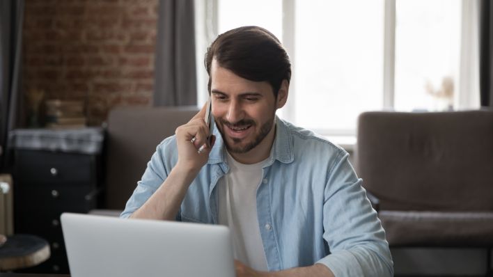 A millennial employee talking on a cellphone while working on their laptop in a home office.