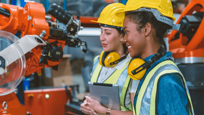Black woman and Asian woman in hard hats interacting with robotic arms in an industrial setting.