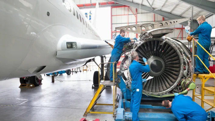 Cuatro hombres con overoles de trabajo azules están trabajando en un motor de avión. El cuerpo del avión es visible a la izquierda. 