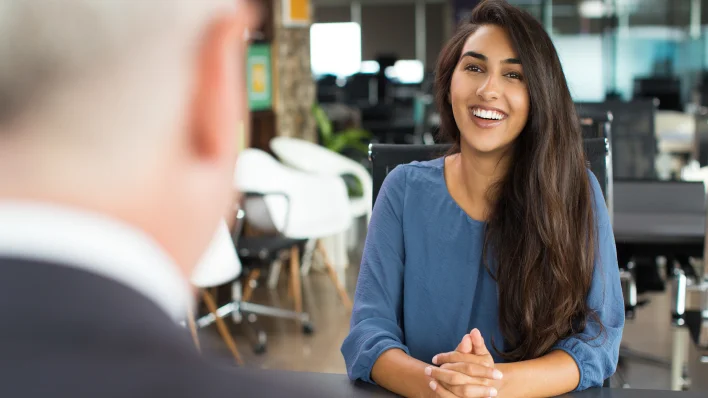 Una joven con cabello largo y castaño y piel morena está sonriendo y haciendo contacto visual con un entrevistador masculino (de espaldas a la cámara). 
