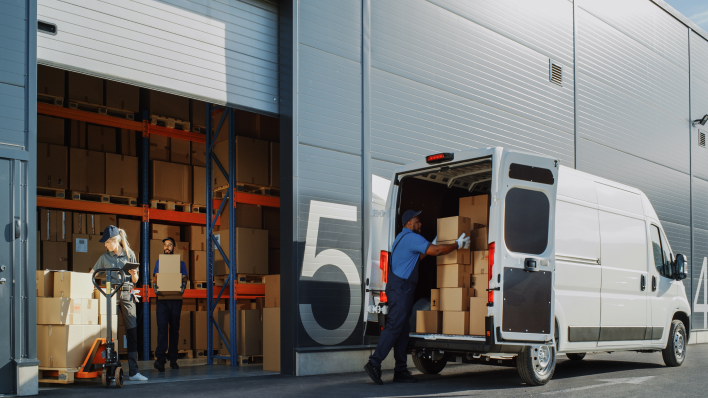 Long shot of man loading boxes into a nearly full white van in front of a loading dock door. We can see a large warehouse interior with another man carrying a large box toward the door.