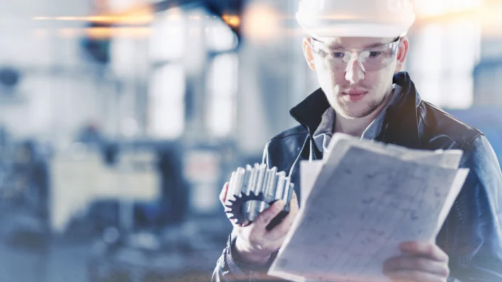 Young man in hard hat and safety goggles holding an industrial part and looking at a blueprint or plan.