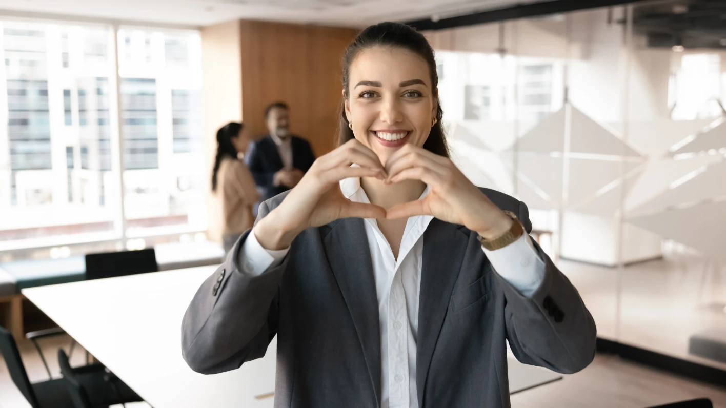 As a result of career planning, this young white woman in a gray business suit is happy; she forms a heart with her hands.