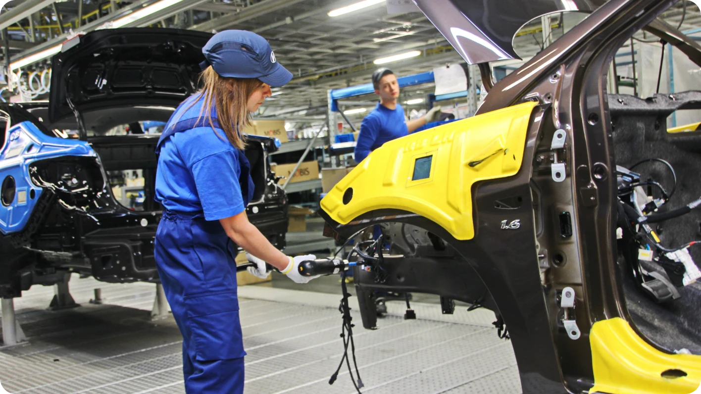 Female automotive worker in blue coveralls is welding a part to a partially assembled car in a factory setting.