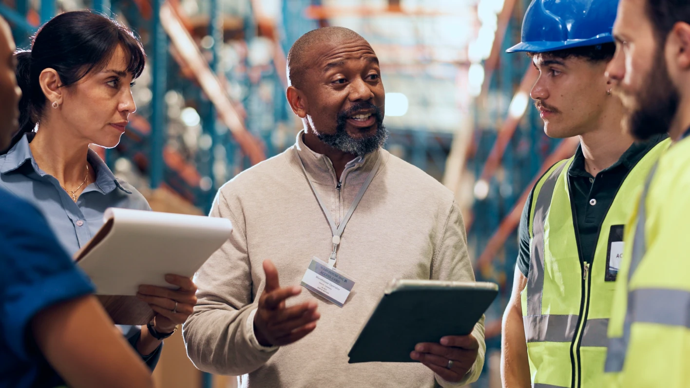 Man with an ID lanyard and beige sweater is holding a tablet and giving instruction to four employees in a factory setting.