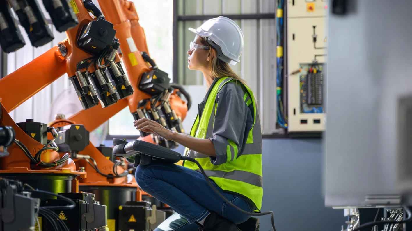 Una mujer con equipo de seguridad controla una máquina robótica con una consola de mano, representando la automatización en la manufactura.