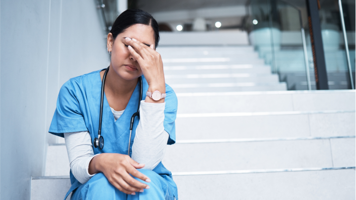 Burnout in healthcare: a woman in medical scrubs sits on a staircase rubbing her eyes. She wears a stethoscope around the neck.