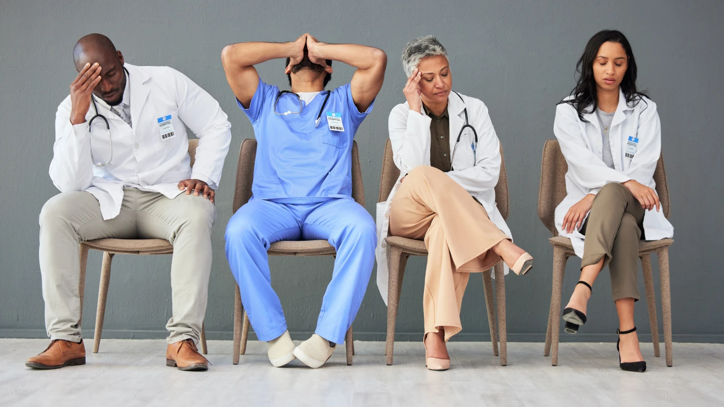 Four tired multi-ethnic healthcare workers in smocks and scrubs sit side-by-side in waiting room-style chairs.