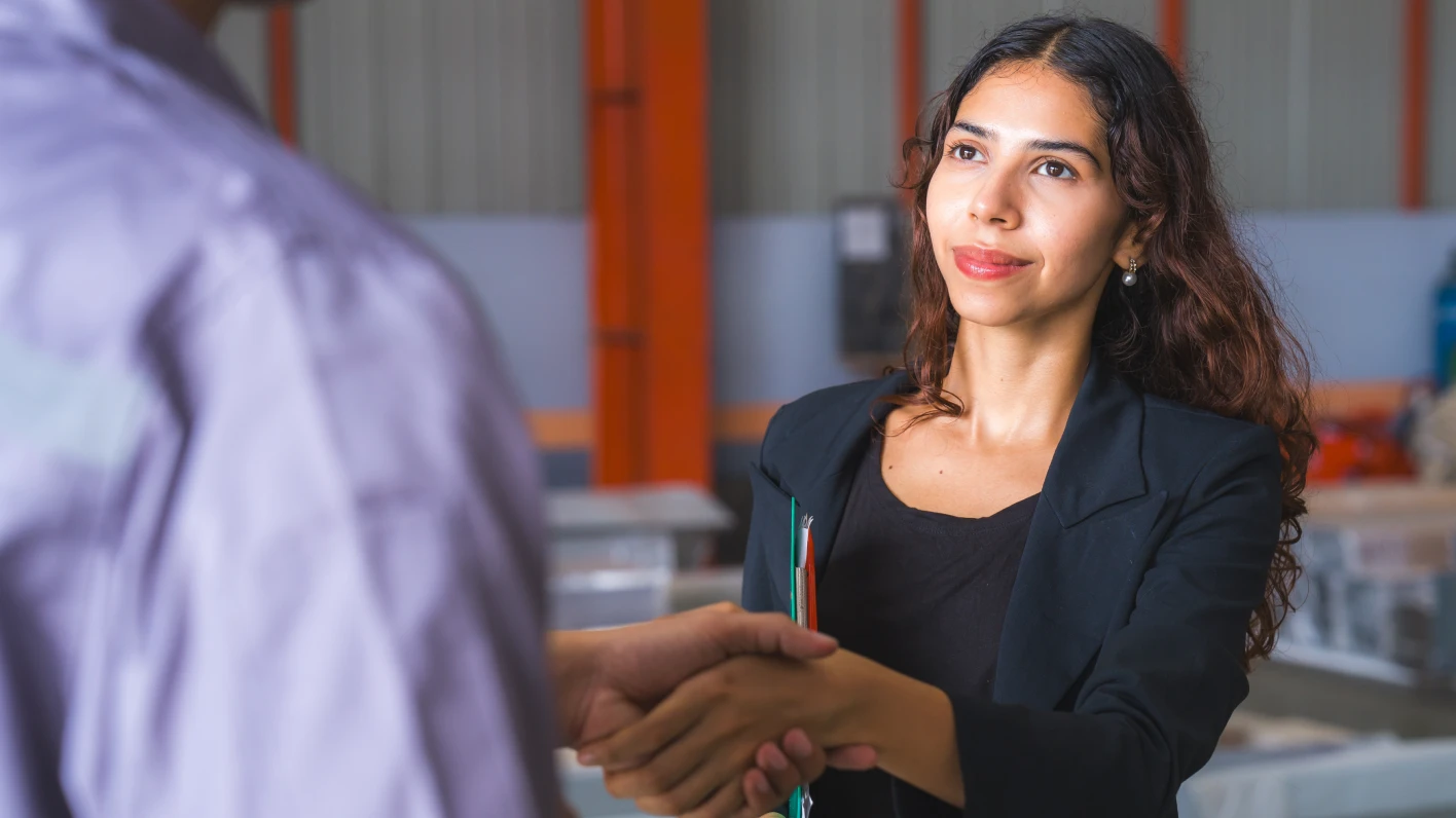 How to nail an interview: a young woman with brown hair and skin shakes hands with an unseen man. She wears a blazer and knit top. 
