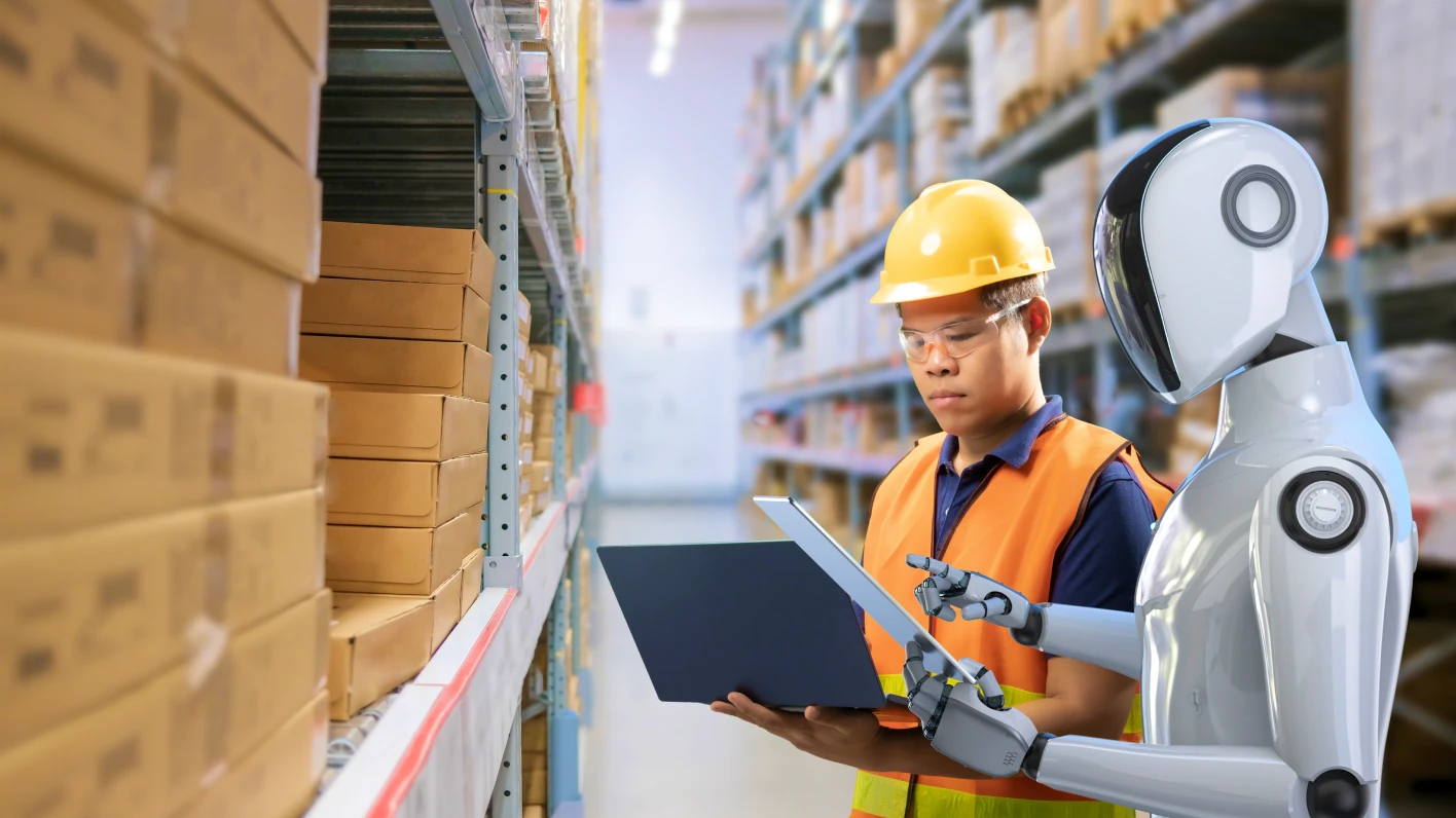 Male order filler in a warehouse is standing next to a robot holding a laptop while the robot holds a tablet.