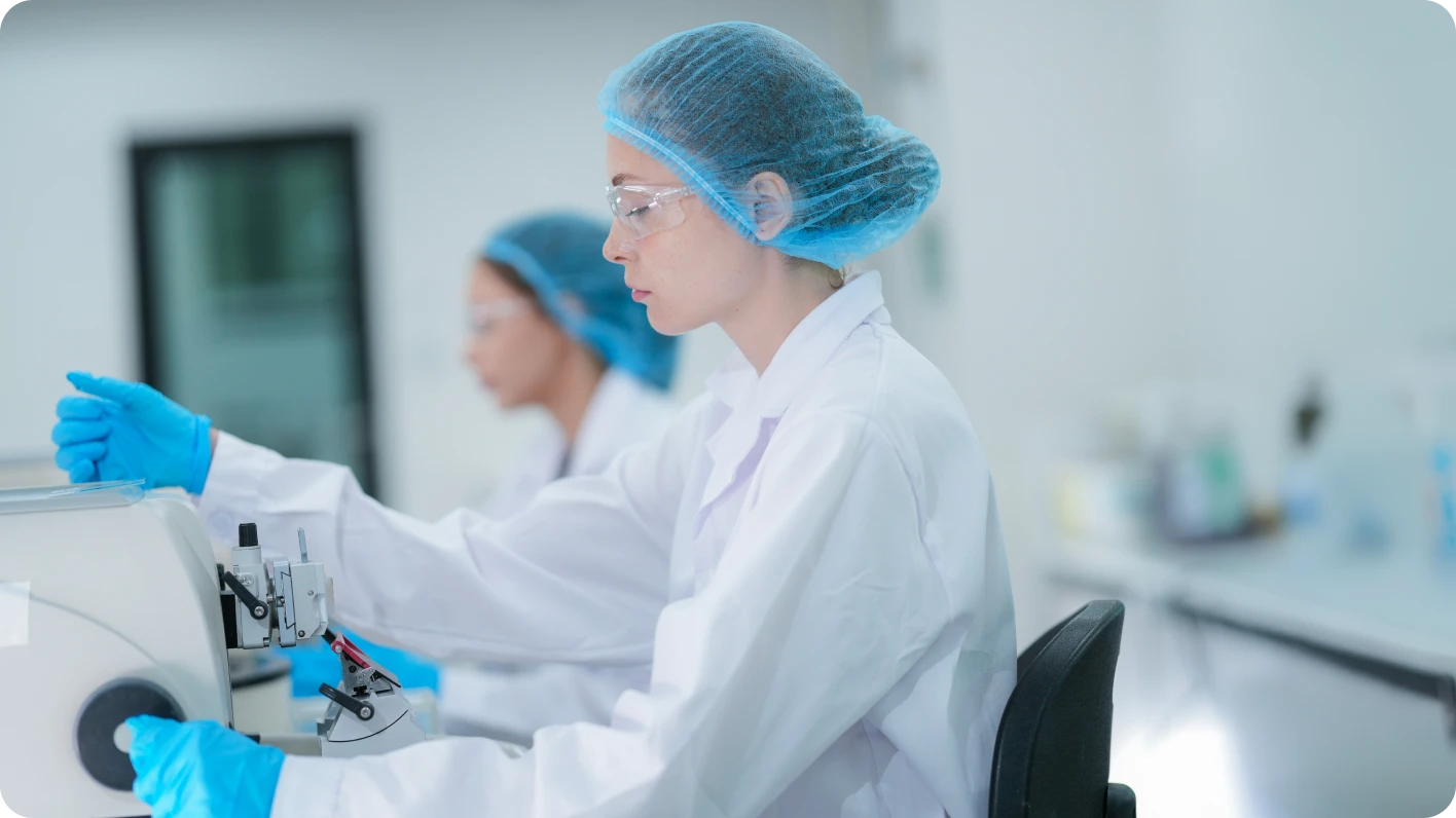 Técnica de laboratorio médico con bata blanca, guantes azules y gorro azul trabajando con una máquina de mesa.