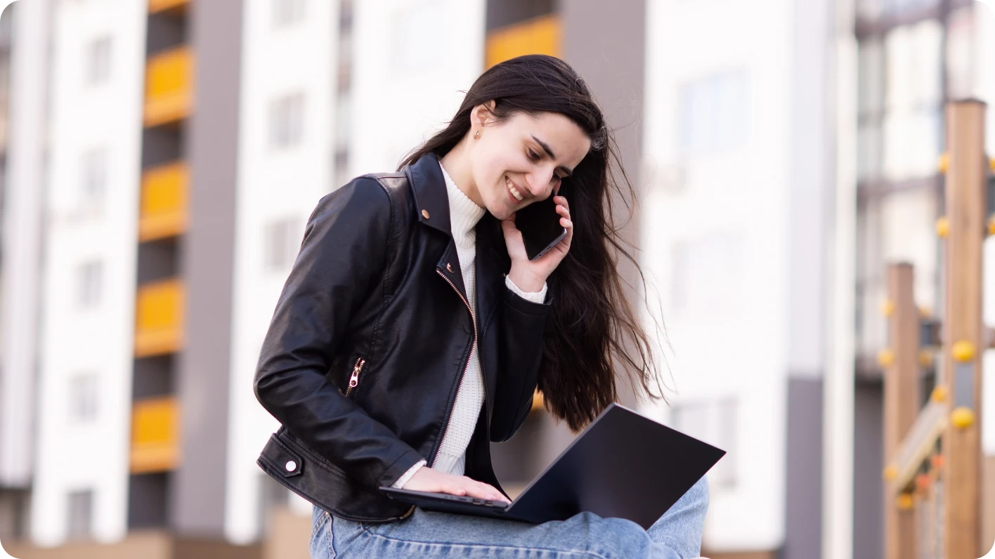 Mujer joven blanca con jeans y chaqueta de cuero se sienta con una laptop en su regazo y un celular en su oído, explorando cómo usar LinkedIn.