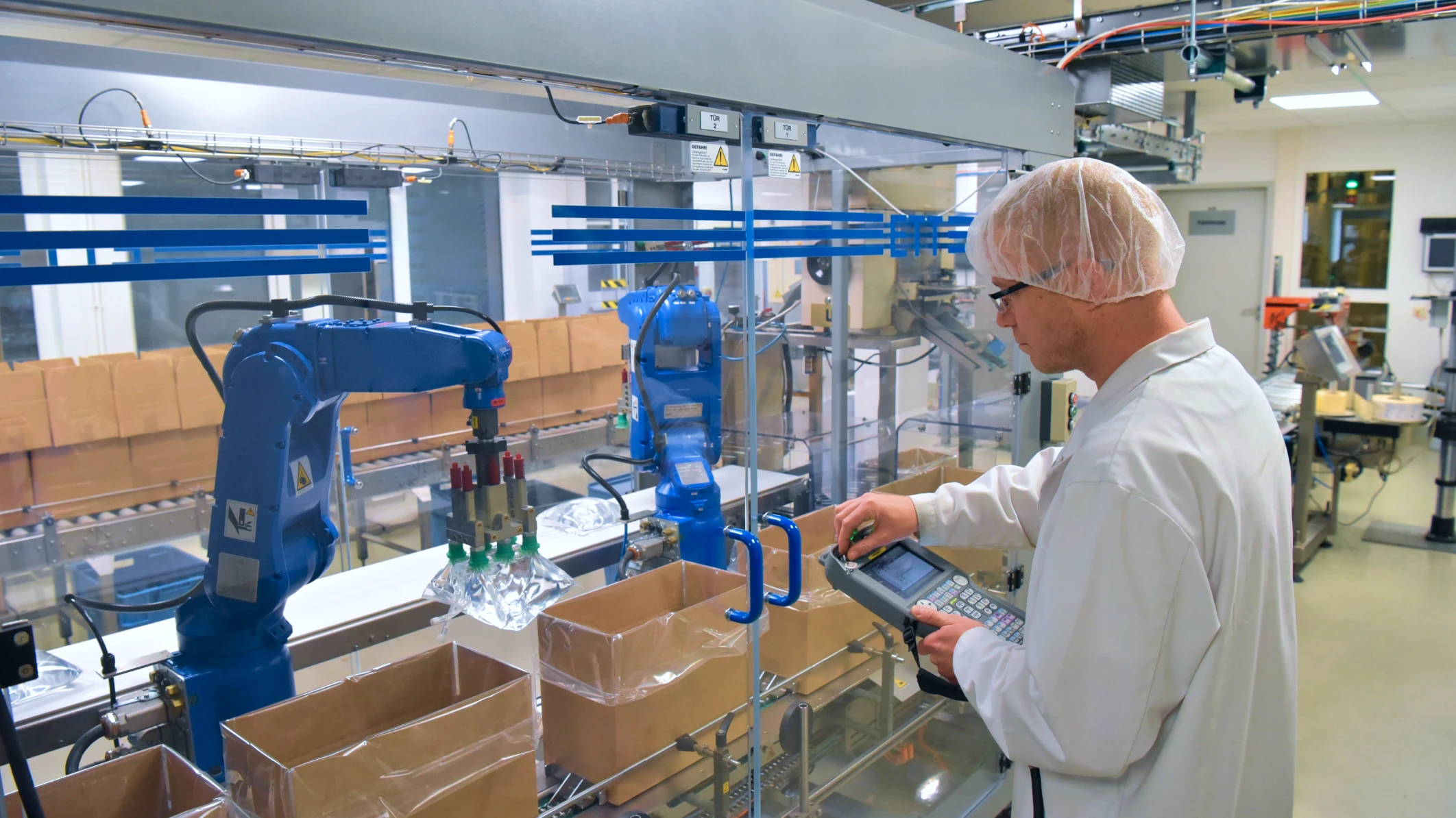 White male in white lab coat and hairnet interacts with a robotic arm in medical device factory setting. He holds a tablet device.