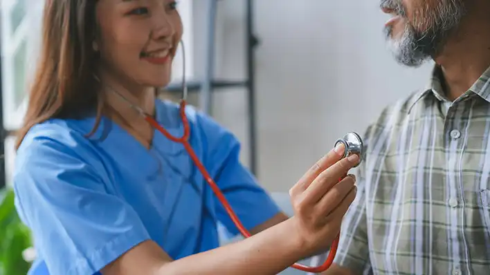A young white female medical assistant holds a stethoscope to the chest of an older bearded man in plaid shirt.