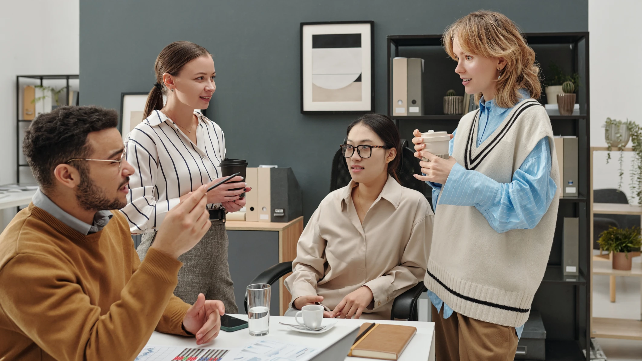 Four young professionals, dressed in business casual attire, stand around a table in an office for an impromptu meeting.
