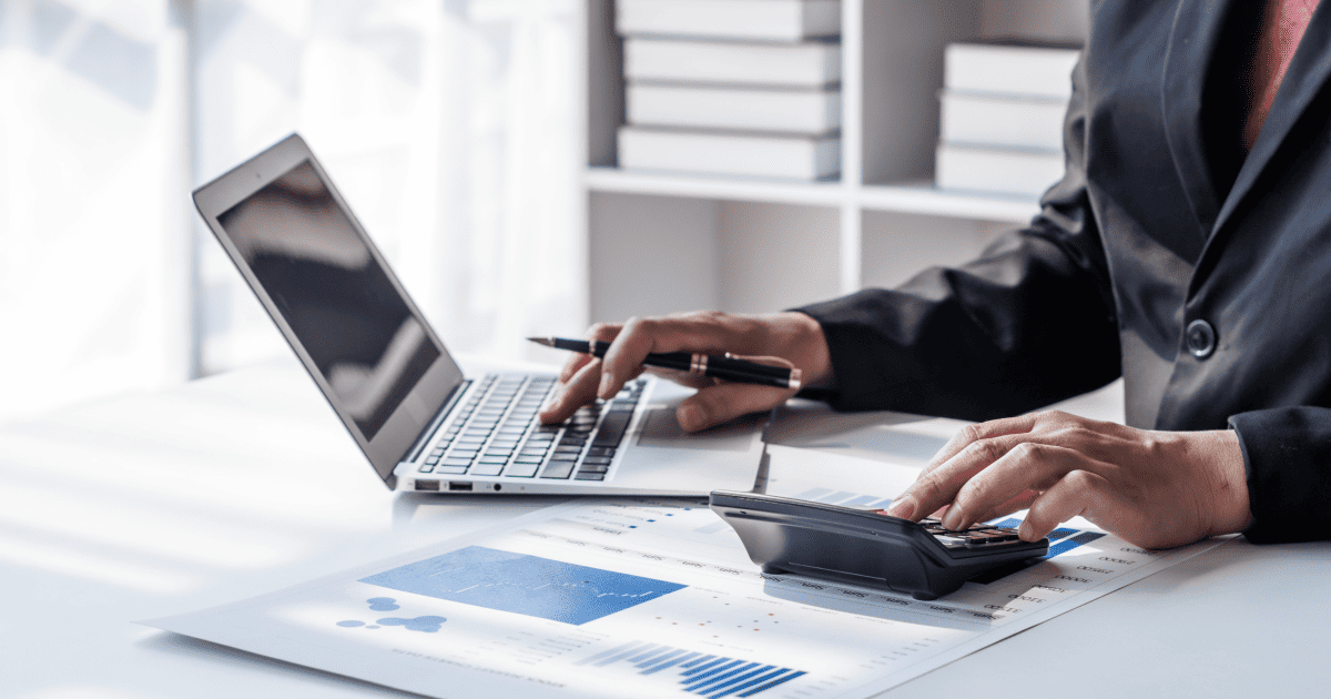 Desktop shot of female figure in dark suit jacket from neck down. She is typing on a laptop with one hand and a calculator with the other. Bar and graph charts are on her desk. 