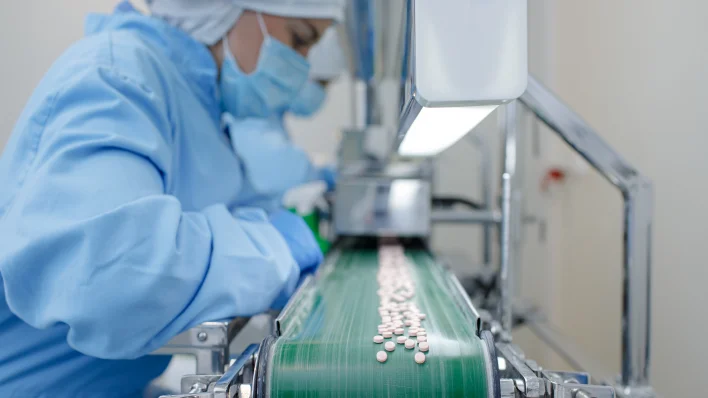 Entorno de producción farmacéutica con una mujer en equipo de protección y mascarilla inspeccionando una línea de montaje llena de pastillas blancas. 