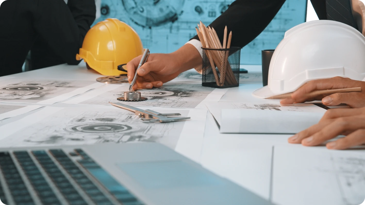 Tabletop featuring architectural drawings, two hard hats, keyboard and two peoples’ hands.
